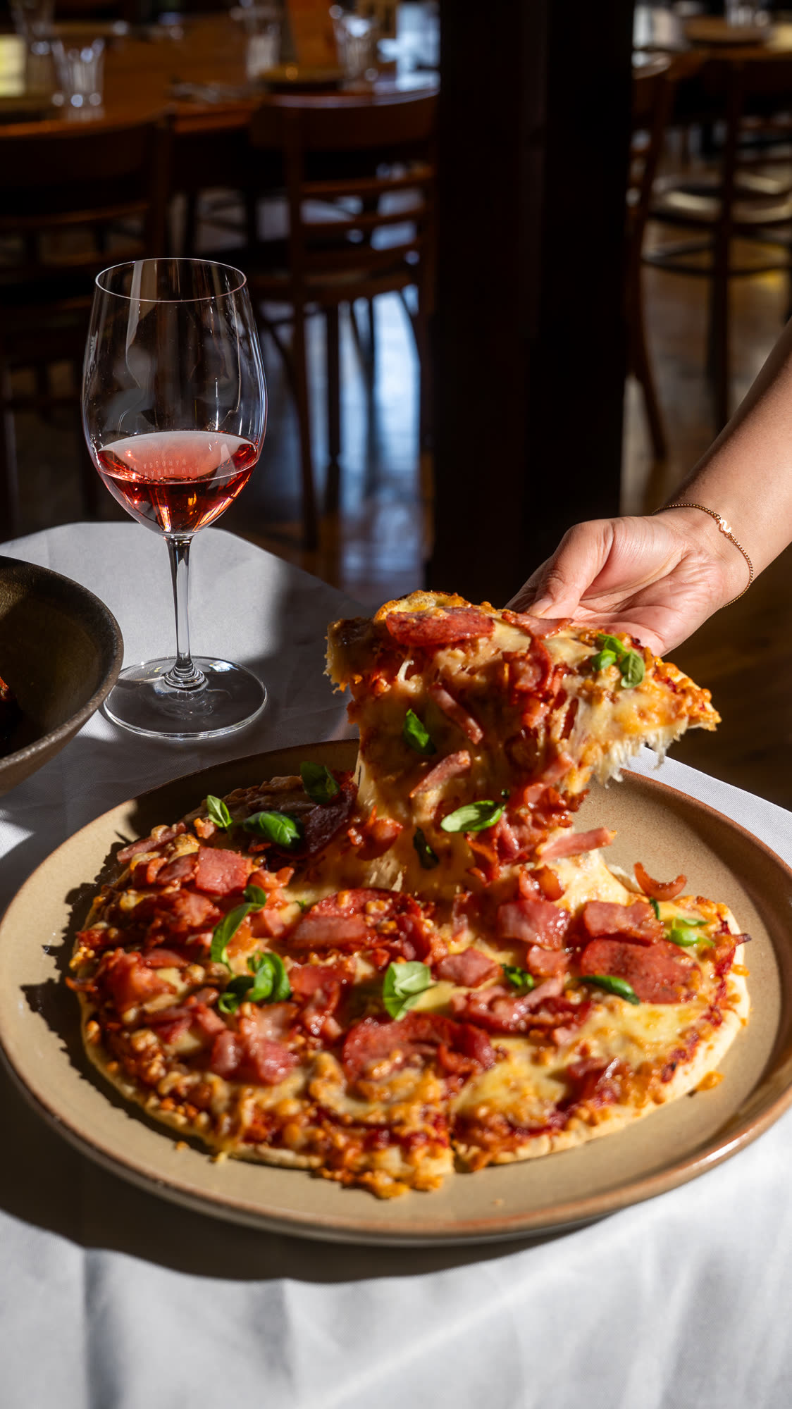 A hand lifting a slice from a shared pizza, with a glass of rosé on the table.