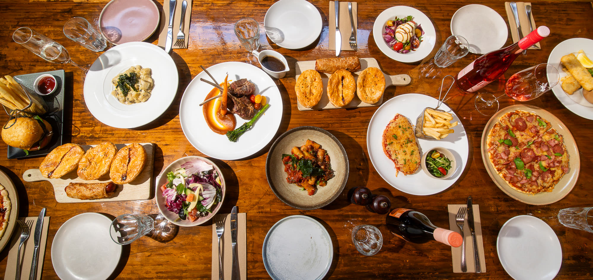 An overhead view of a timber table laid with shared dishes, salads and wine.