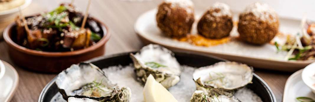 A platter of oysters on ice beside a bowl of braised pork belly and a plate of arancini.