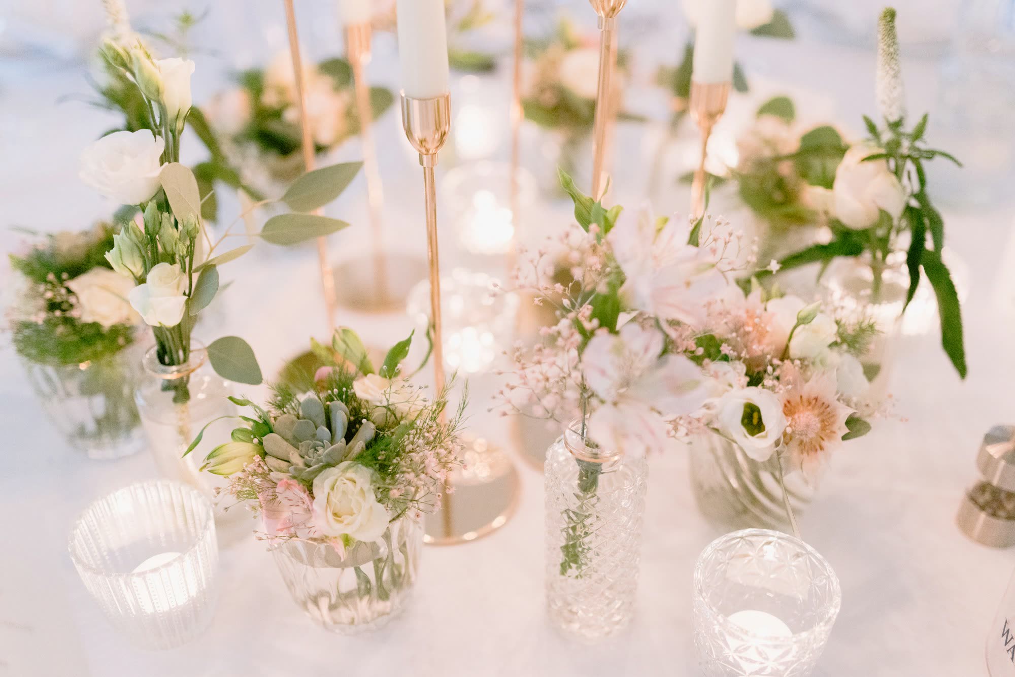 Small bud vases of flowers and tall taper candles on a white tablecloth, generic stock photo pending real venue photography.